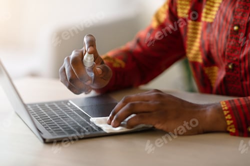 Preview: Unrecognizable african man sanitizing laptop keyboard surface with disinfectant spray, closeup