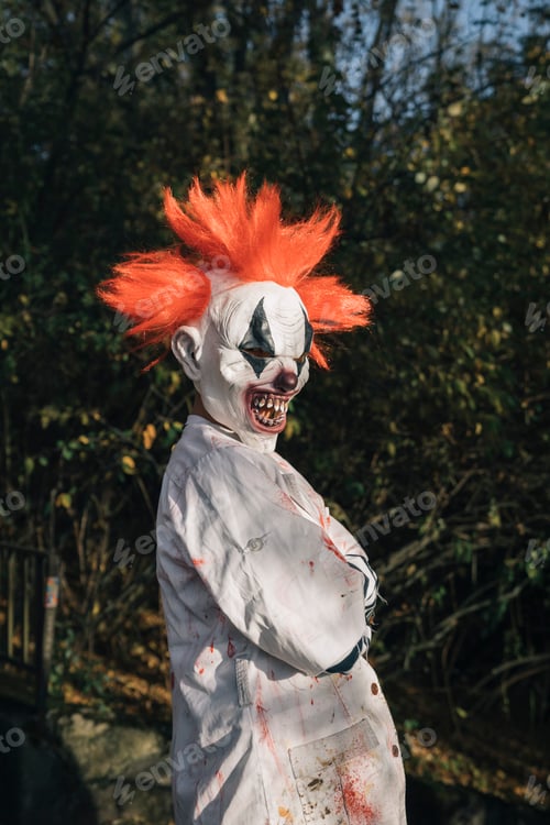 Preview: Halloween scary clown wearing bloody costume standing in forest
