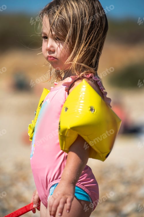 Preview: Young child pouting on a sandy beach with arm floats and a shovel.