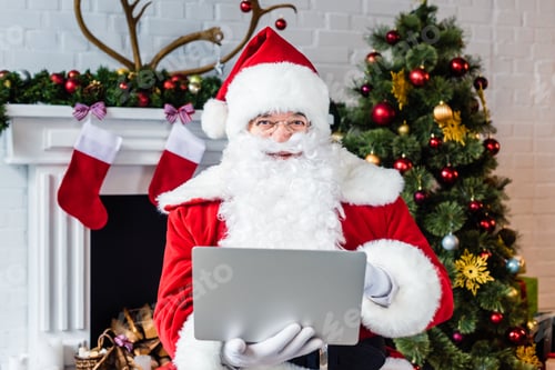 Preview: santa claus using laptop and looking at camera near fireplace