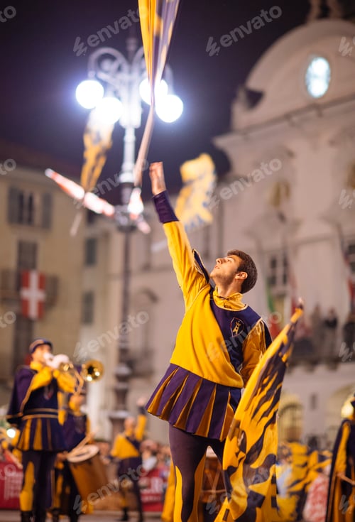 Preview: A flagbearer in vibrant medieval attire performs at a historical festival at night.