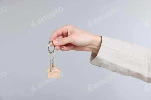 Preview: Real estate agent holding key with house shaped keychain on gray background, closeup