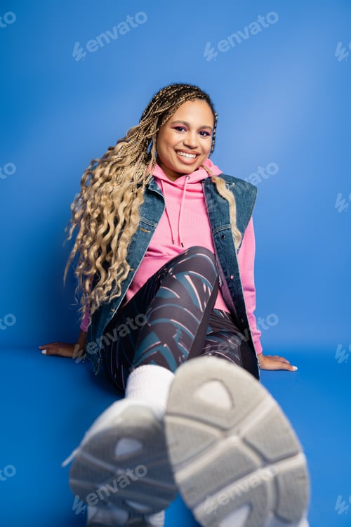 Preview: happy dark skinned woman with dreadlocks sitting in sporty outfit on blue background, look at camera