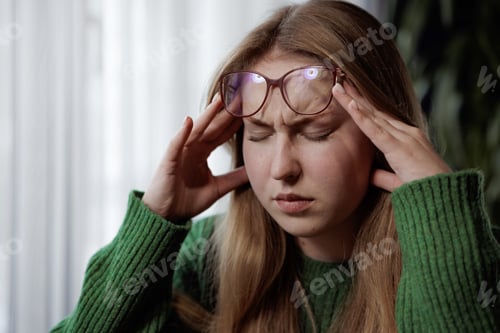 Preview: Young woman with headache touching temples, showing signs of stress or fatigue at home or work.