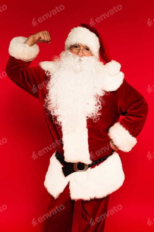 Preview: Santa claus in costume showing muscle on arm on red background