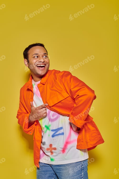 Preview: portrait of excited indian man in orange jacket and diy t-shirt looking away on yellow backdrop