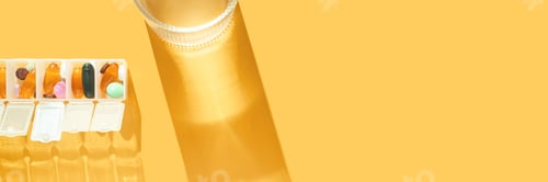 Preview: Assorted pills in weekly organizer and glass of water on bright yellow