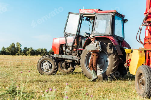 Preview: selective focus of bearded farmer in straw hat using laptop near tractor