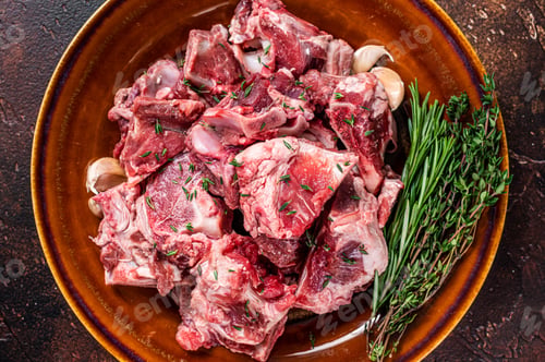 Preview: Raw mutton meat diced for goulash or stew with bone on a rustic plate. Dark background. Top view