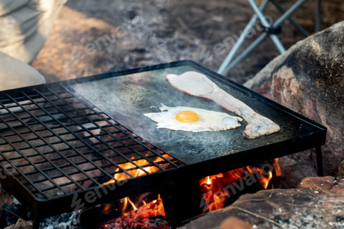 Preview: Breakfast camp cooking. Grilling crispy bacon and eggs on a cast iron bbq plate over the camp fire.