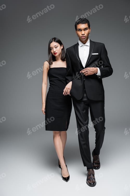 Preview: full length of multiethnic couple in elegant formal wear walking and looking at camera on grey