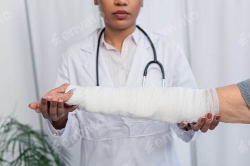 Preview: Cropped view of blurred african american doctor touching hand of elderly woman in plaster bandage