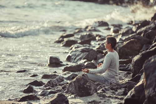 Preview: Candid authentic male millennial sitting in lotus pose on sea rocky shore greets dawn
