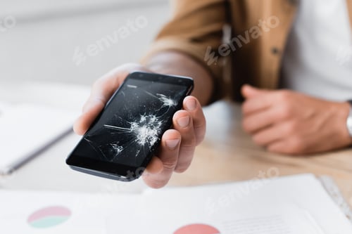 Preview: Cropped view of man holding smashed smartphone with blurred workplace on background