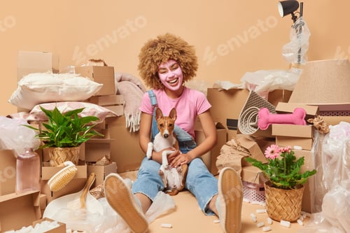 Preview: Positive woman with curly hair applies beauty mask wears t shirt and denim overalls poses with dog p