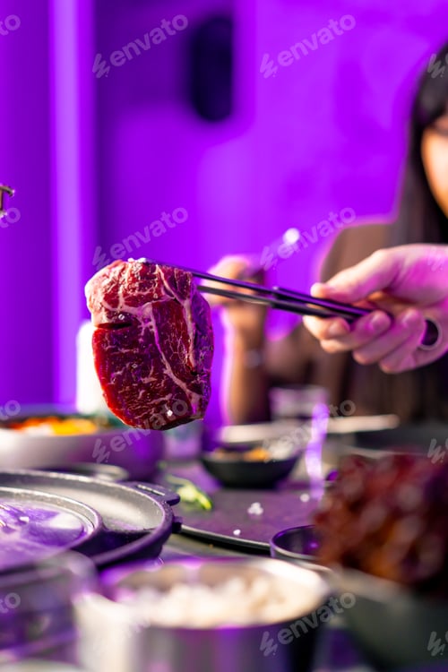 Preview: close-up of a hand with tongs picking up a juicy raw piece of meat to put on the grill in restaurant
