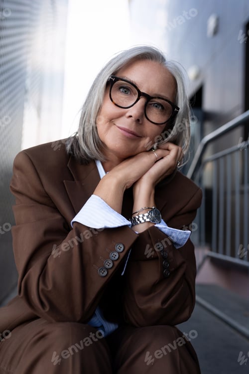 Preview: well-groomed healthy slender gray-haired mature business lady dressed in a stylish brown jacket sits
