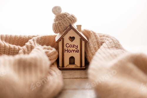 Preview: A small house, wrapped in a scarf with a hat, stands on a wooden background. Heating system concept