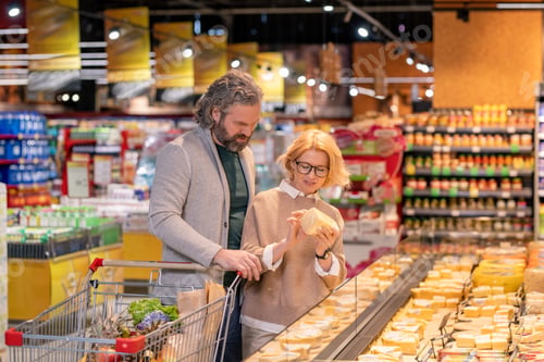 Preview: Blond mature woman in eyeglasses looking at price on piece of cheese