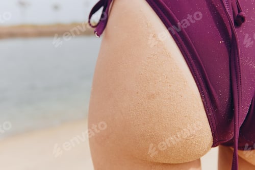 Preview: Close up of woman's buttocks in purple swimsuit on beach near water with another body of water in