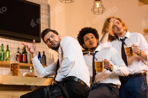 Preview: emotional young multiethnic businessmen drinking beer and watching match in bar