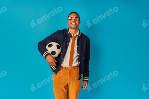 Preview: pleased african american student in orange pants, with soccer ball, smiling at camera on blue