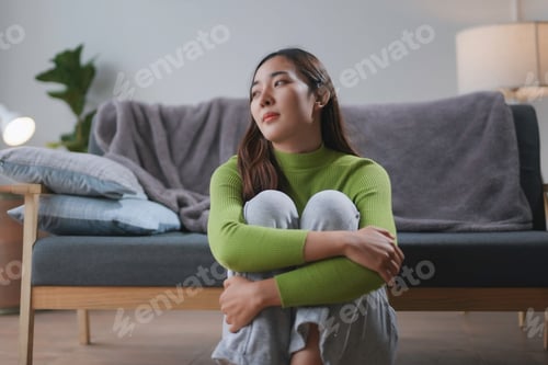 Preview: Young woman is sitting alone on the floor at home, hugging her knees and looking away with a sad
