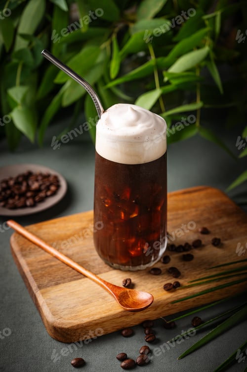Preview: Iced coffee brew with foam in a glass on a wooden board on a green background with coffee beans
