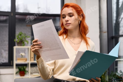 Preview: Young woman with red hair reviews documents at her modern office workspace