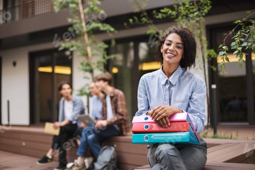 Preview: Pretty smiling lady sitting on bench with colorful folders on knees and happily looking in camera