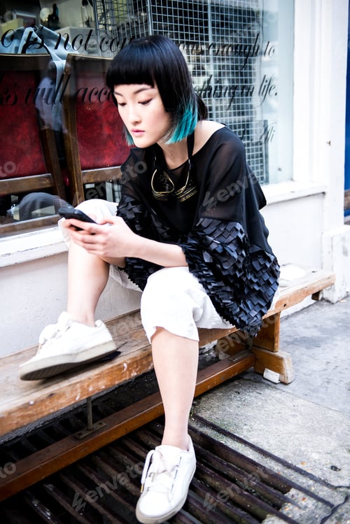 Preview: Young stylish woman waiting outside shop looking at smartphone