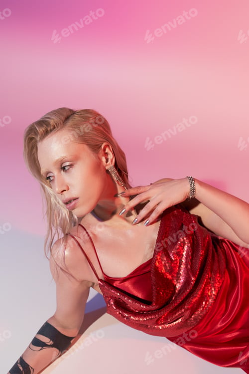 Preview: appealing woman with accessories and tattoo in red chic dress reclining on floor on pink backdrop