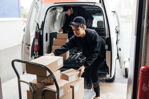 Preview: Two people in uniform loads parcels in van. Woman taking boxes from cart.