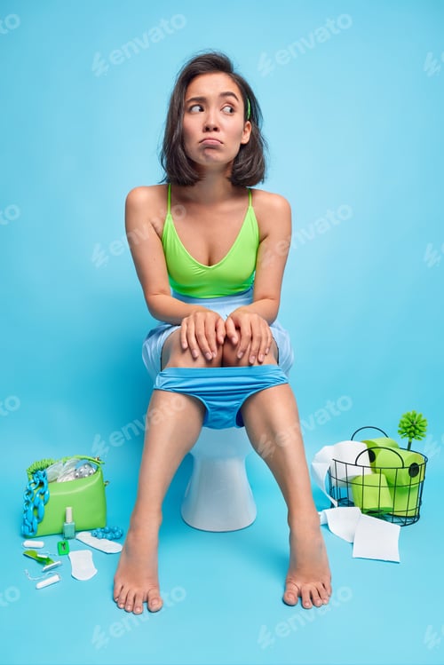 Preview: Thoughtful unhappy Asian woman sits on toilet bowl looks displeased aside keeps hands on knees poses