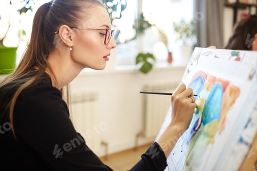 Preview: Young beautiful girl in glasses dressed in black blouse sits at the easel and paints a picture in
