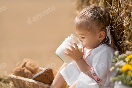 Preview: Happy cute little baby in folk clothes sits on a wheat field with a jar of milk