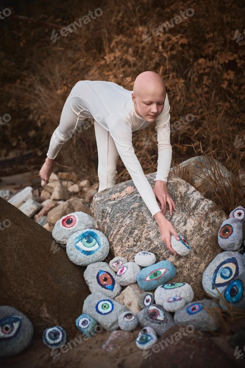 Preview: Young hairless girl with alopecia in white futuristic costume looking at surreal lot of rocks eyes