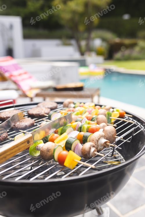 Preview: A barbecue grill is cooking skewers with vegetables and meat by a poolside with copy space