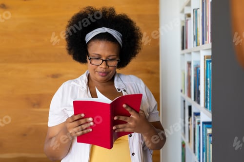 Preview: Happy plus size african american woman wearing glasses, standing by bookshelf looking at book