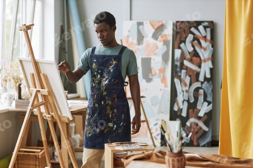 Preview: Young serious man in denim apron standing in front of canvas on easel