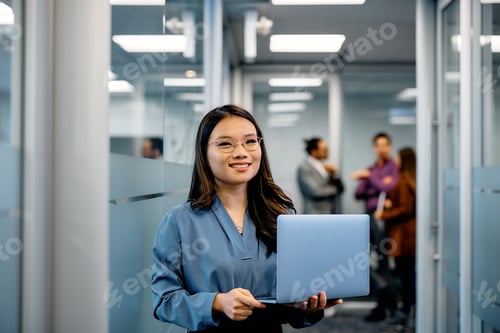 Preview: Happy Asian female entrepreneur with laptop in a hallway looking at camera.