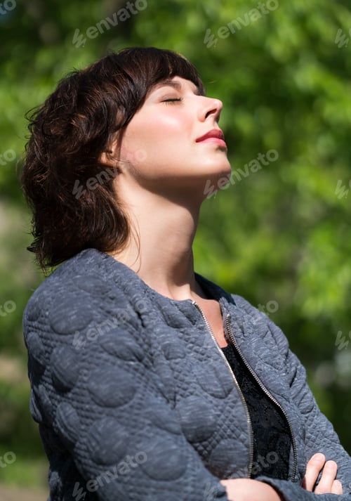 Preview: Beautiful young woman enjoying the sunlight outdoors, eyes closed with background of green leaves.