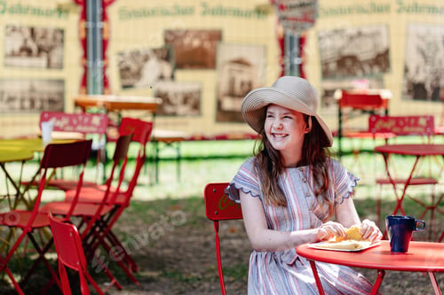 Preview: A girl in a hat and a dress eating food in a street cafe at a summer fair