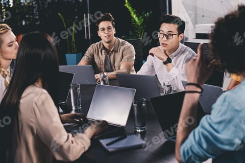 Preview: multicultural group of business partners having discussion at table with laptops in modern office