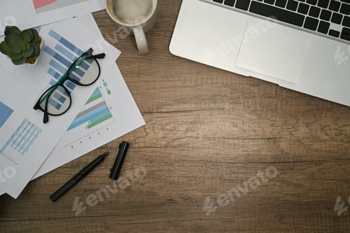 Preview: Above view of laptop, glasses, coffee cup and financial documents on wooden table.