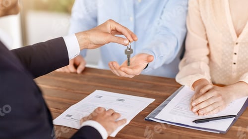 Preview: Real estate agent giving house key to young couple after signing property agreement at table, close