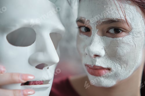 Preview: Teenage girl with a clay cosmetic mask applied to her face holds a white theater mask in hand.