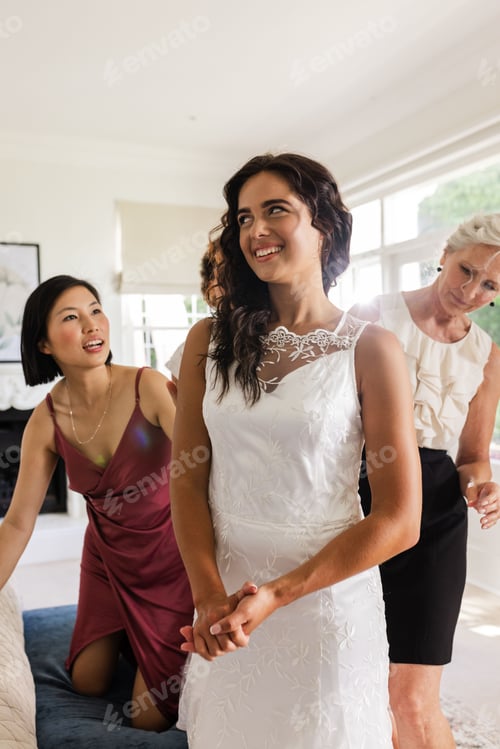 Preview: Bride in elegant white dress smiling with bridesmaids helping at home