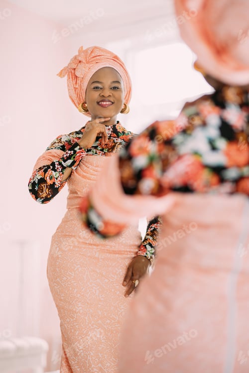 Preview: Pretty African woman in elegant fashionable pink colorful dress and turban, looking at herself at