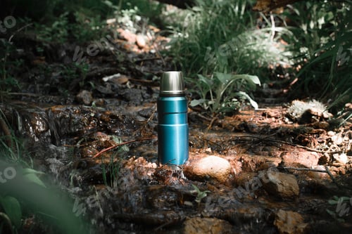 Preview: Thermos standing in water outdoors, forest and stream on a summer day. Hiking flask with a hot drink